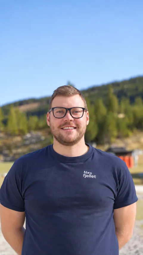 Portrett av en smilende mann med briller i mørk T-skjorte, med skogkledd fjell og blå himmel i bakgrunnen.